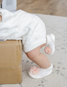 Baby wearing a white knit romper and flower-accented white booties, standing on a patterned rug and leaning on a cardboard box.