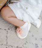 A baby's leg and foot wearing a white cable-knit dress and a white slipper sock adorned with a pink flower.