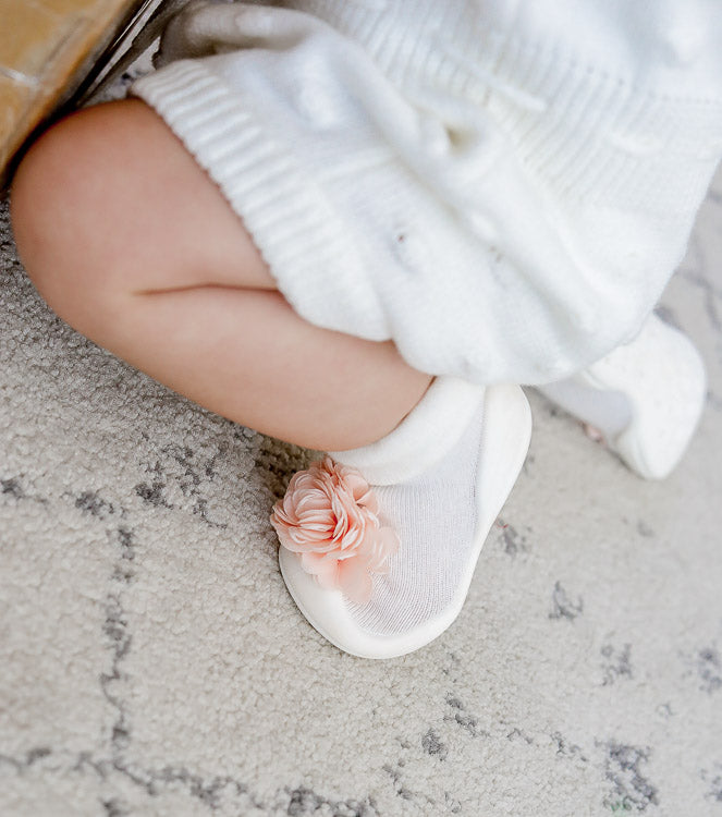 A baby's leg and foot wearing a white cable-knit dress and a white slipper sock adorned with a pink flower.