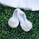 Two white baby shoes with white fabric flower embellishments on a green plant background.