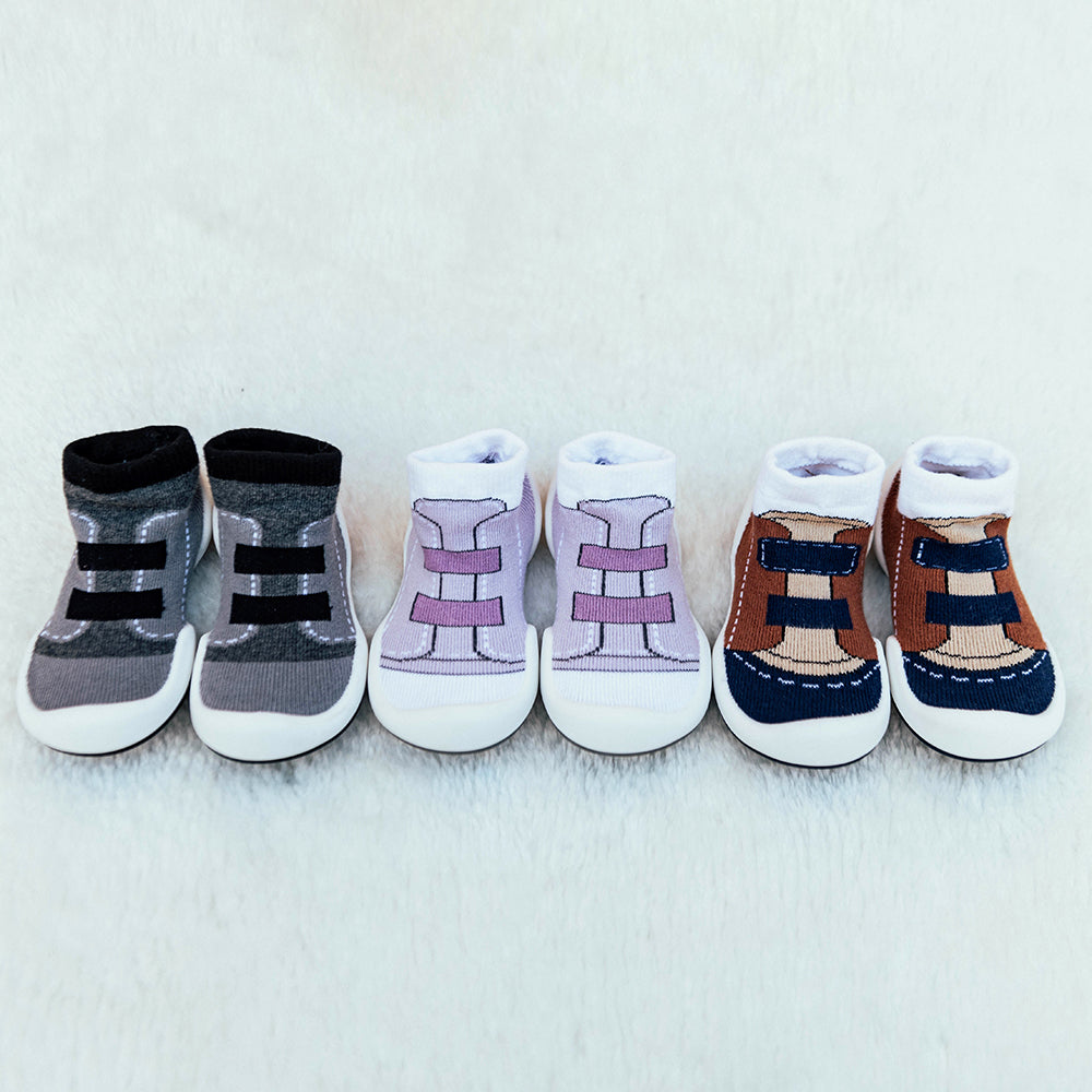 Three pairs of toddler shoe-socks in gray, lavender, and brown, aligned on white fabric.