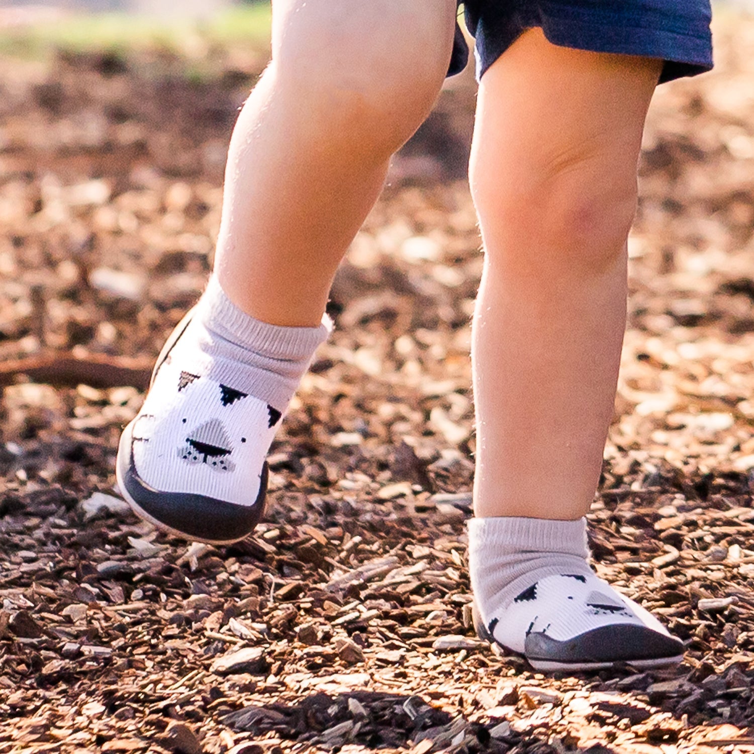 Child's legs wearing gray sock shoes with a bear design on wood chips.