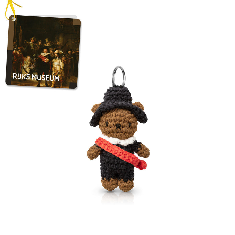 Crocheted teddy bear in a black outfit with a red sash, wearing a black hat. Keyring at the top. Ruks Museum tag in the background.