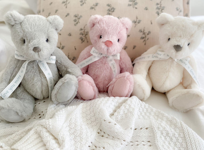 Three teddy bears in gray, pink, and white sitting on a textured surface.