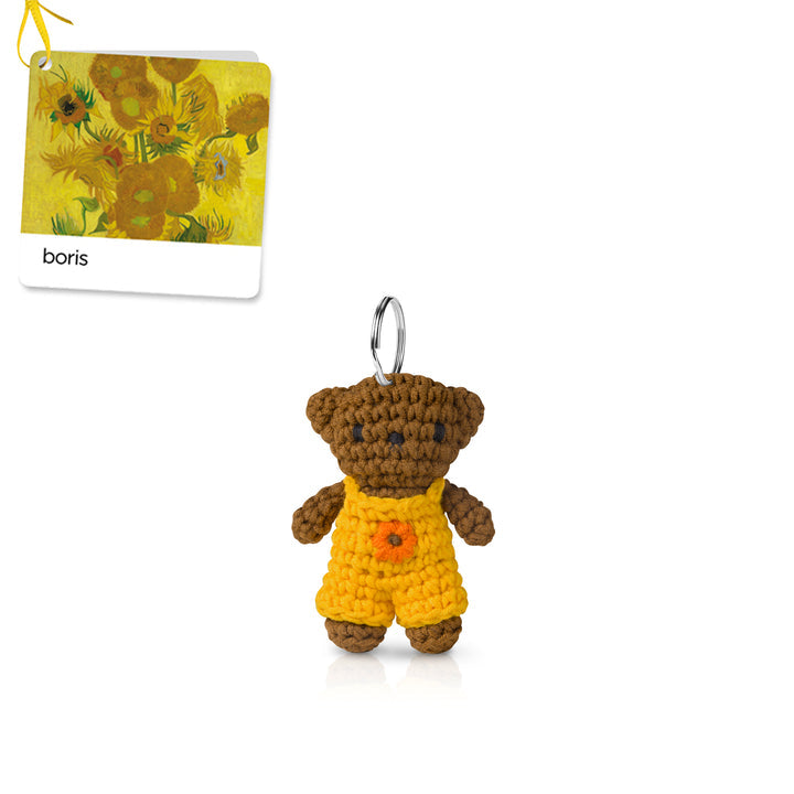 Crocheted brown teddy bear keychain in yellow overalls with a flower.
