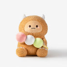 A plush toy character with a fuzzy tan body, small white horns, and a simple face, holding three soft balls in pink, white, and light green, photographed on a clean white background.