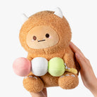 A plush toy character with a fuzzy tan-brown body, small white horns, and a simple face, being held by two hands against a clean white background. The plush is holding three round pastel items: pink, white, and light green.