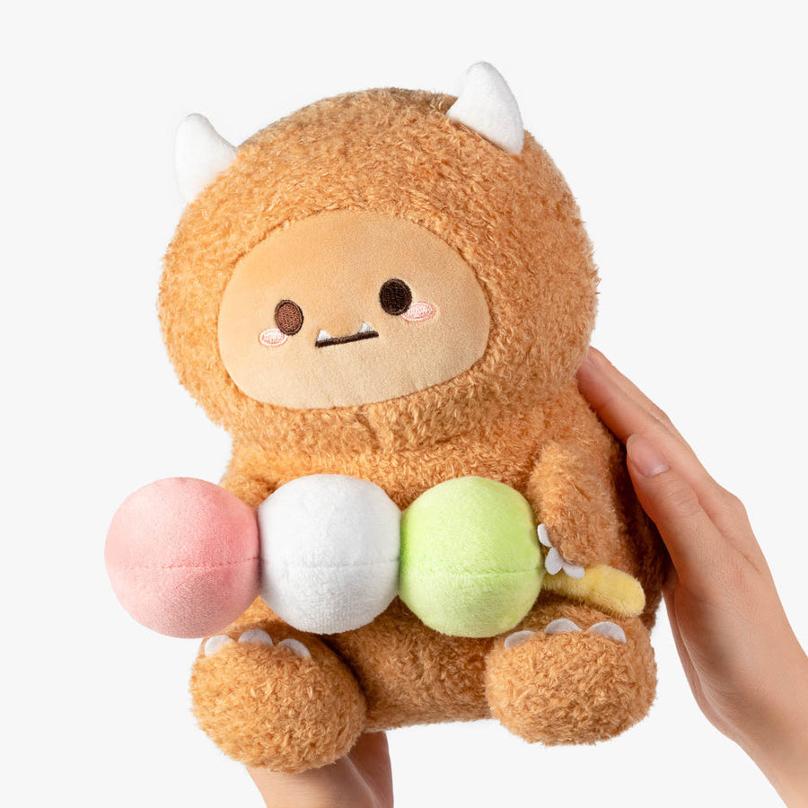 A plush toy character with a fuzzy tan-brown body, small white horns, and a simple face, being held by two hands against a clean white background. The plush is holding three round pastel items: pink, white, and light green.