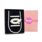 A pearl and crystal jewelry set displayed in an open black gift box next to its pink branded box. The set includes a long pearl strand, a pearl and crystal collar with a cream bow, and several crystal and pearl hair clips.