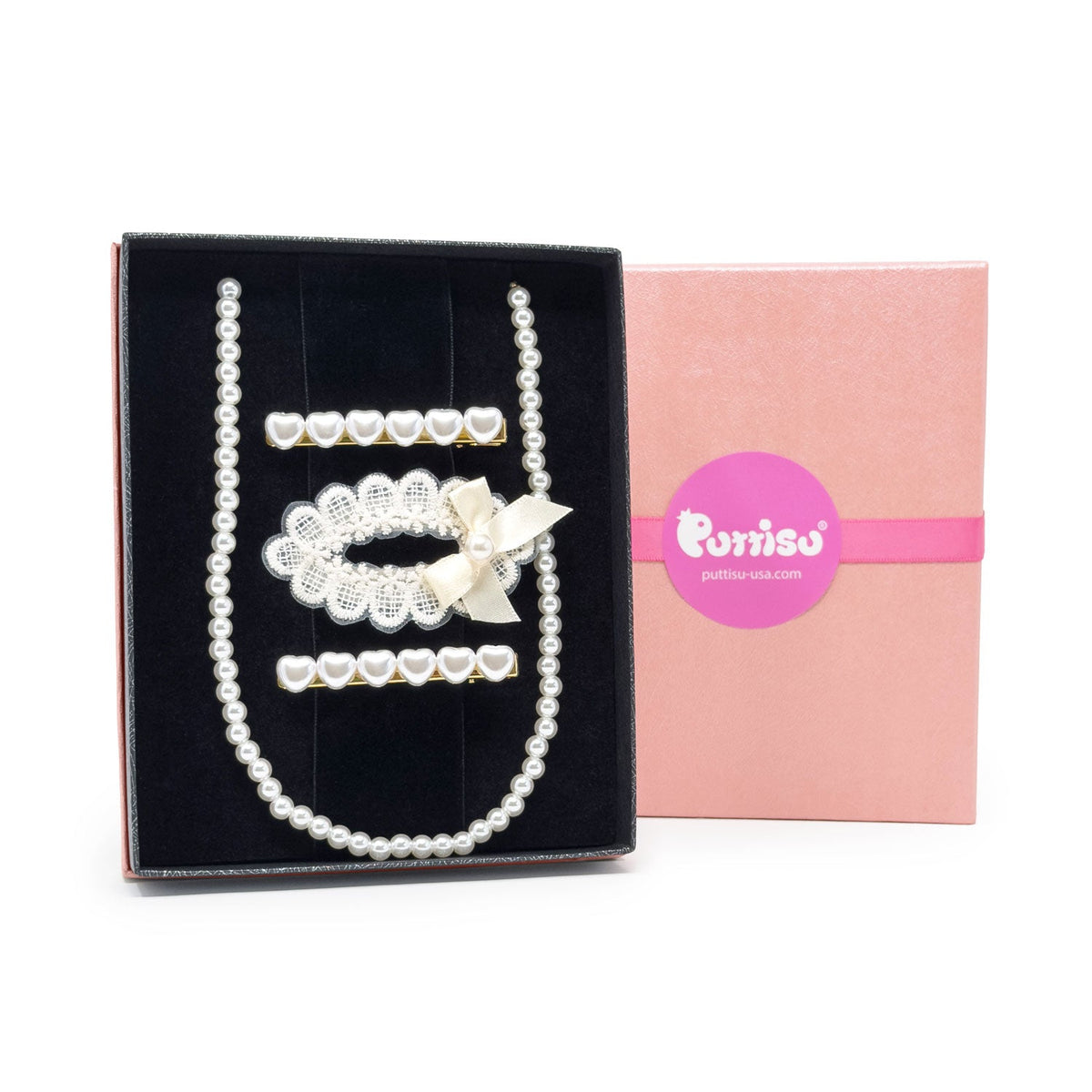 A pearl and crystal jewelry set displayed in an open black gift box next to its pink branded box. The set includes a long pearl strand, a pearl and crystal collar with a cream bow, and several crystal and pearl hair clips.