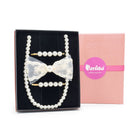 A pearl hair accessory set displayed in a black presentation box next to its pink branded box. The set includes a long pearl strand, two pearl hair clips with gold-tone ends, and a white lace bow with a central pearl.