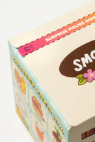 Close-up of a collectible toy box showing a pink flap sticker that reads 'SURPRISE FIGURE INSIDE' and part of the brand name 'SMO' on a cream-colored box with pastel illustrations.