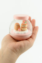 Hand holding a small glass container with decorative food-themed figures on a white background