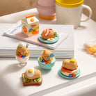 A set of cute cartoon breakfast-themed miniature figurines displayed on a white tabletop next to a pink and yellow mug, with a notebook and a small milk carton in the background.