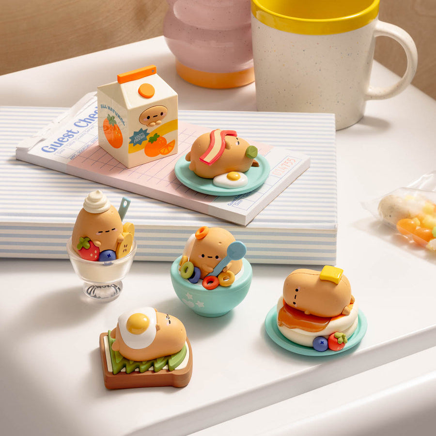 A set of cute cartoon breakfast-themed miniature figurines displayed on a white tabletop next to a pink and yellow mug, with a notebook and a small milk carton in the background.