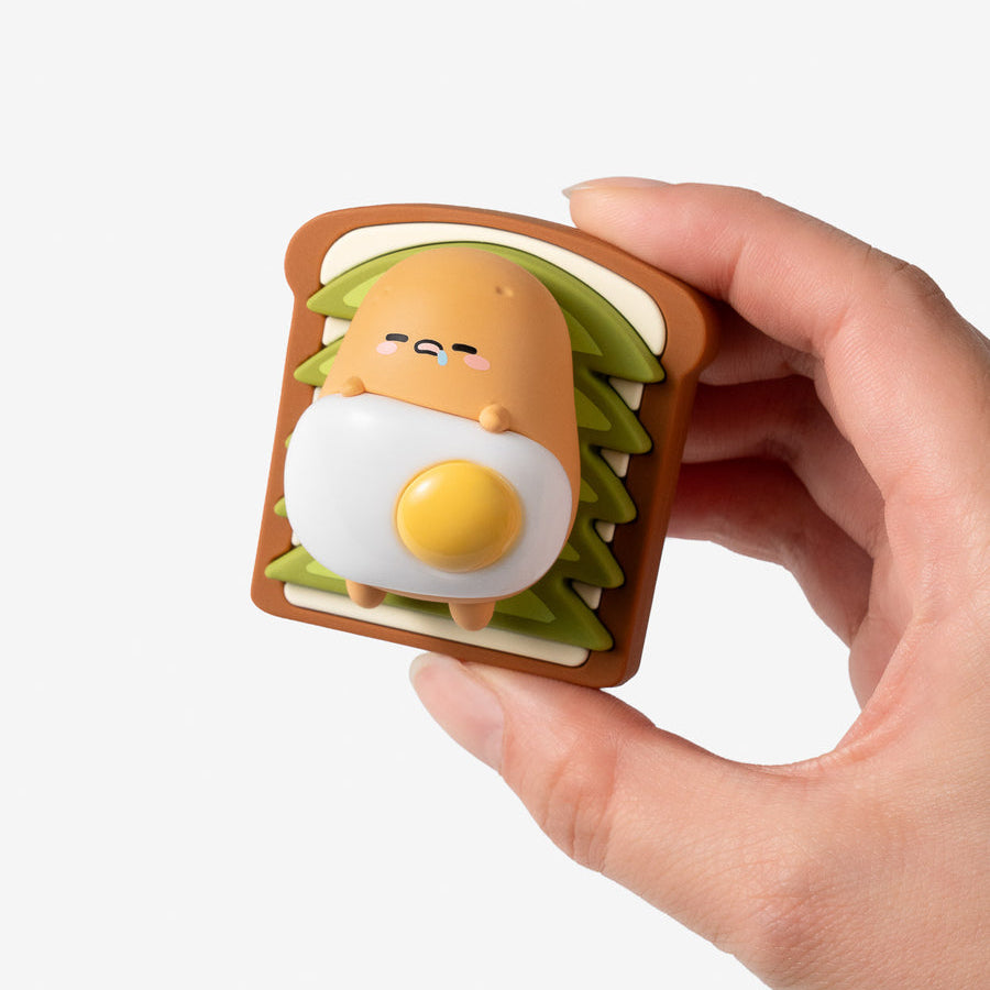 A hand holding a small, cute, egg-shaped character figurine designed to look like it is inside a slice of toast with green lettuce and white cream.