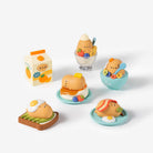 Five small cute breakfast-themed figurines and a milk carton set on a clean white background. The characters have round orange-brown faces and are shown with eggs, toast, a sandwich, a cup with a spoon, and a milk carton.