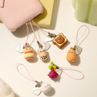 A set of cute, small 3D cartoon charms with pink cords, displayed on a white surface next to a pink case, a yellow book, and a green candle. The charms include a hamburger, a bubble tea cup, a slice of toast with a bear, a sleeping cat, a pink dinosaur, and a white cat face.