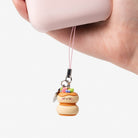 A hand holding a light pink phone case with a dangling charm. The charm is a small character-shaped macaron with a colorful sprinkle topping and a silver metal ring.
