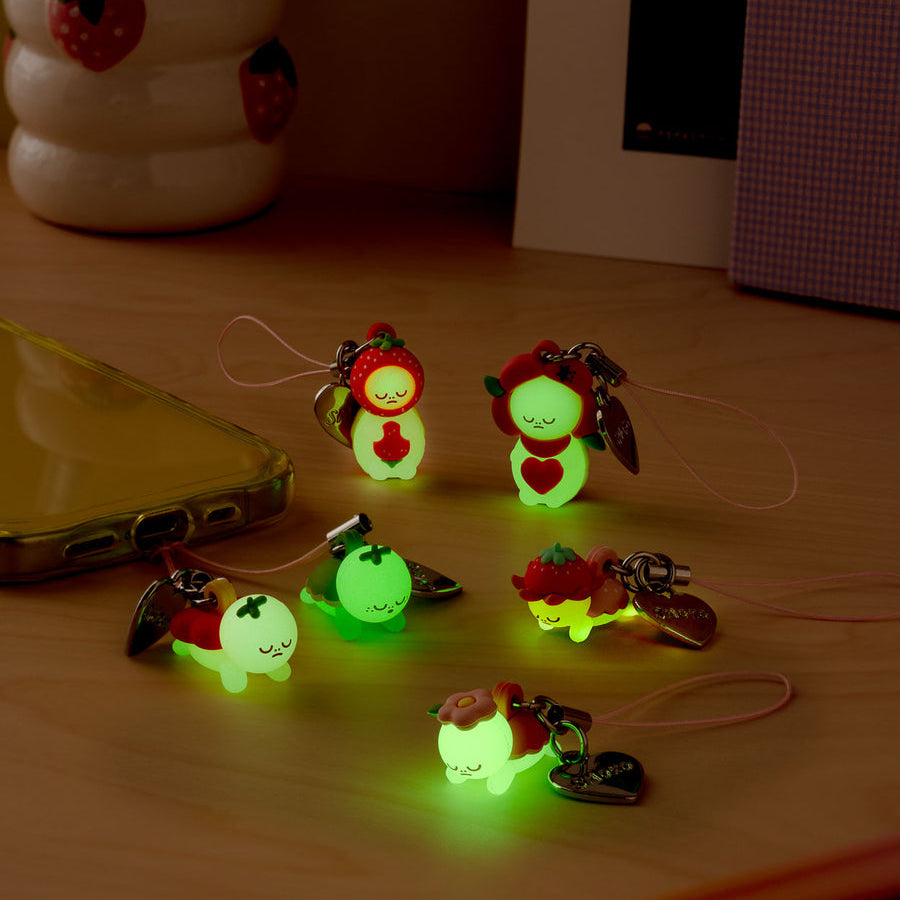 Several small glowing fairy-shaped keychains with red and green strawberry-themed accessories are arranged on a wooden desk in a dimly lit room, with a cup, phone, and pencil visible in the background.