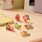 A set of cute, small cartoon phone strap charms with metal keychains, displayed on a light wood desk next to a yellow phone case, a pink pen, and a strawberry-themed cup.