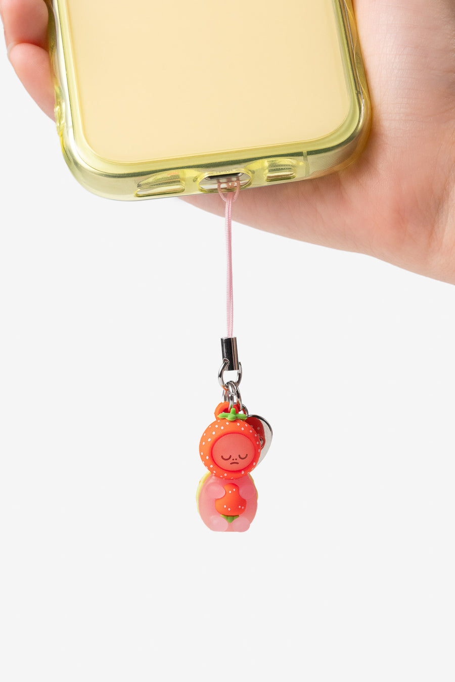 A close-up of a hand holding a phone with a translucent pale yellow case, attached to a pink wrist strap and a small strawberry-shaped charm.