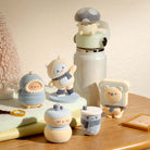 A collection of cute, pastel-colored character figurines and accessories arranged on a wooden table next to a white water bottle. The characters feature soft blue, beige, and gray tones, with one wearing a mushroom cap and another shaped like a bear on toast.