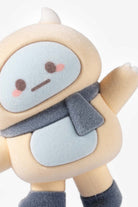 A close-up of a soft plush toy with a round beige body, a pale blue face panel, a dark gray scarf, and dark gray boot covers, shown against a light gray background.