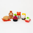 Six small, cute, food-themed figurines arranged on a seamless white background. The set includes a brown character with an orange headband on an orange base, a dark red berry cupcake with green leaves on a striped base, an orange cat in a striped box, a white character with a horn in a black-and-white base, a green-topped white character in a red base, and a tan character lying on an orange base.