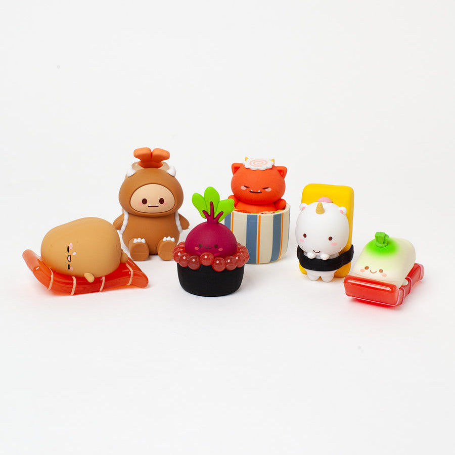 Six small, cute, food-themed figurines arranged on a seamless white background. The set includes a brown character with an orange headband on an orange base, a dark red berry cupcake with green leaves on a striped base, an orange cat in a striped box, a white character with a horn in a black-and-white base, a green-topped white character in a red base, and a tan character lying on an orange base.