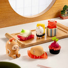 A set of cute, stylized food-themed figurines resembling sushi and snacks, arranged on a light wood tray and tabletop next to a bowl of rice and chopsticks. The figures have a soft, plush or rubbery texture and are decorated with various colorful toppings and wrappers.