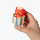 A hand holding a small decorative figurine shaped like a red cat sitting inside a striped cup filled with yellow items, against a plain white background.