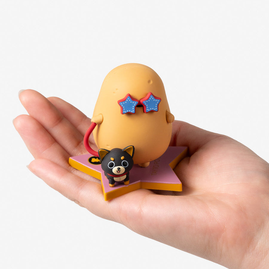 A hand holding a small yellow egg-shaped figurine with blue star-shaped sunglasses and a small black-and-white cat figure on a pink and yellow star base, against a light gray background.
