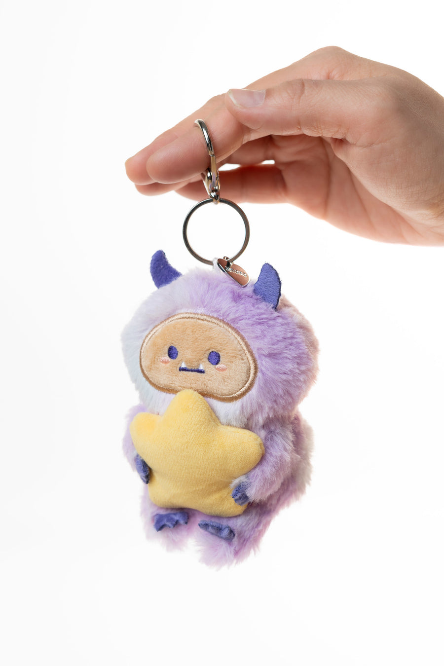 Plush keychain shaped like a purple creature with blue accents holding a yellow star, held by a hand against a white background.