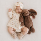 Brown plush bunny toy with long floppy ears and soft fur, shown with baby in floral dress and knitted booties on textured white blanket, lifestyle view