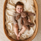 Brown plush bunny toy with long floppy ears and soft fur, held by a baby in a woven basket with cream knit blanket, top-down lifestyle view