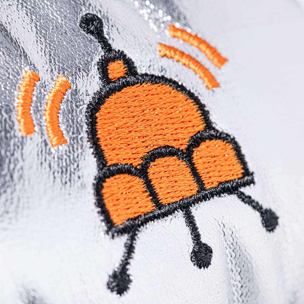 Orange and black embroidered jellysaucer design on silver glittery fabric, by Jellycat, close-up detail showing antennae and signal waves