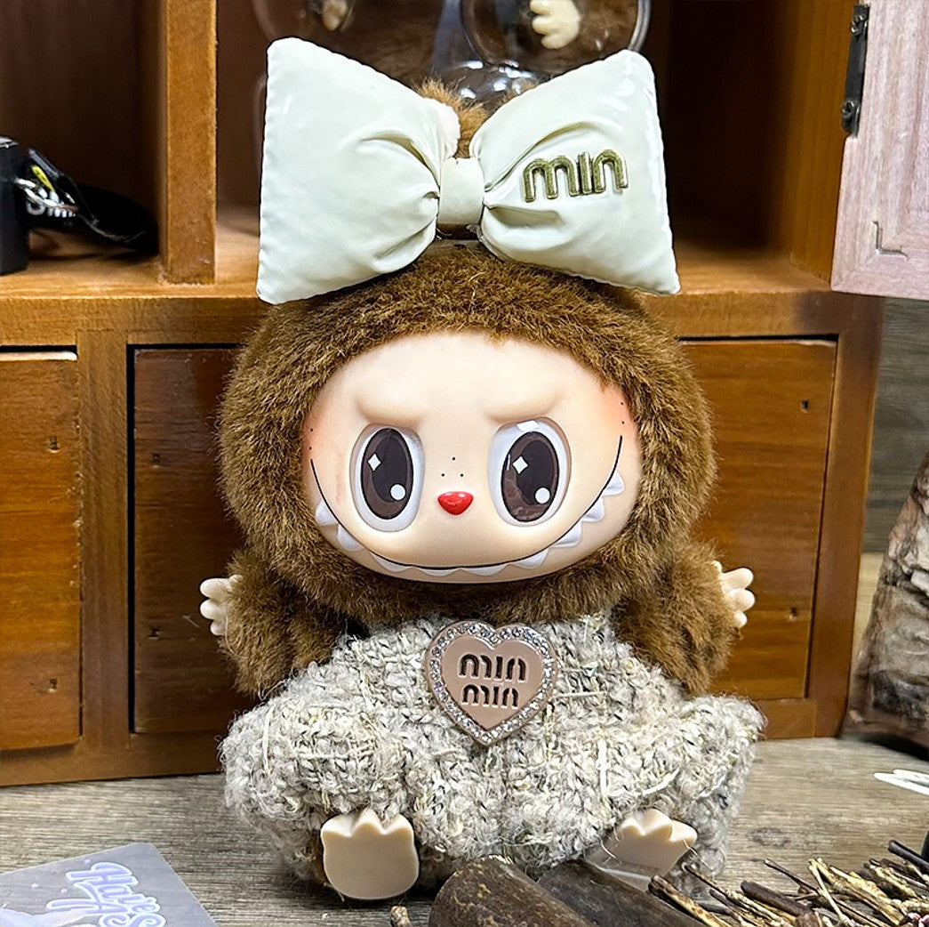 A doll with brown fur, a beige bow, and a textured dress, sitting on a wooden surface.

