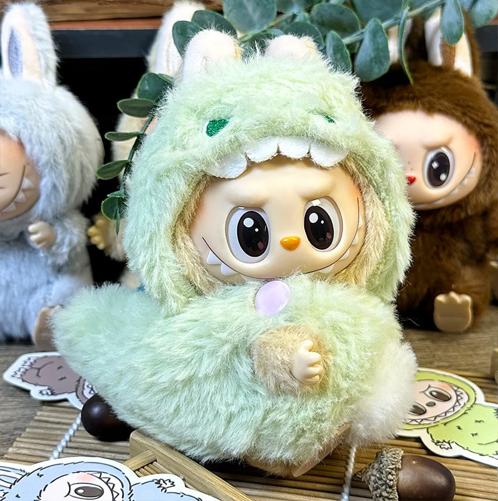 Close-up of three plush dolls. The doll in the center wears a fuzzy, green dinosaur costume. The dolls beside it wear fuzzy gray and brown bunny costumes. The dolls are on a wooden surface with stickers and acorns.