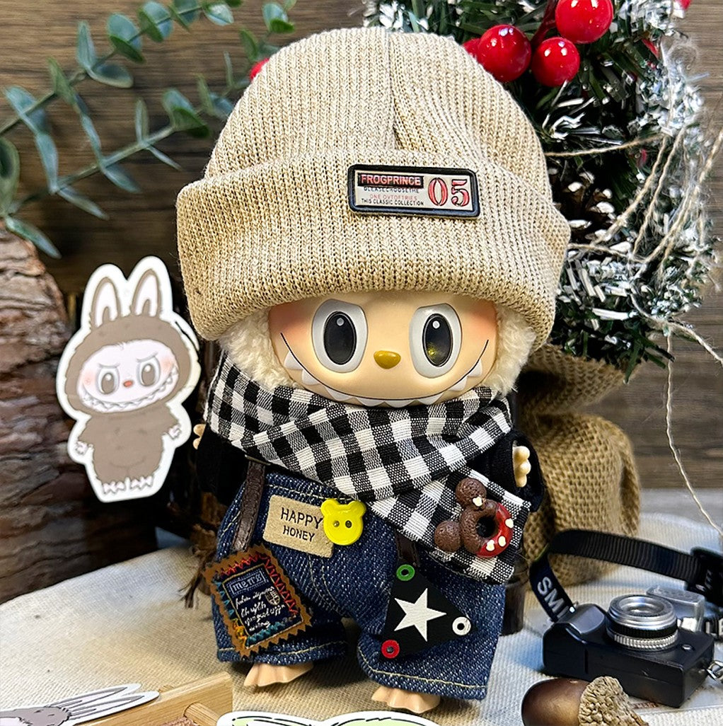 A stylized doll with a tan beanie, a black-and-white checkered scarf, denim overalls with playful patches, standing against a festive backdrop.
