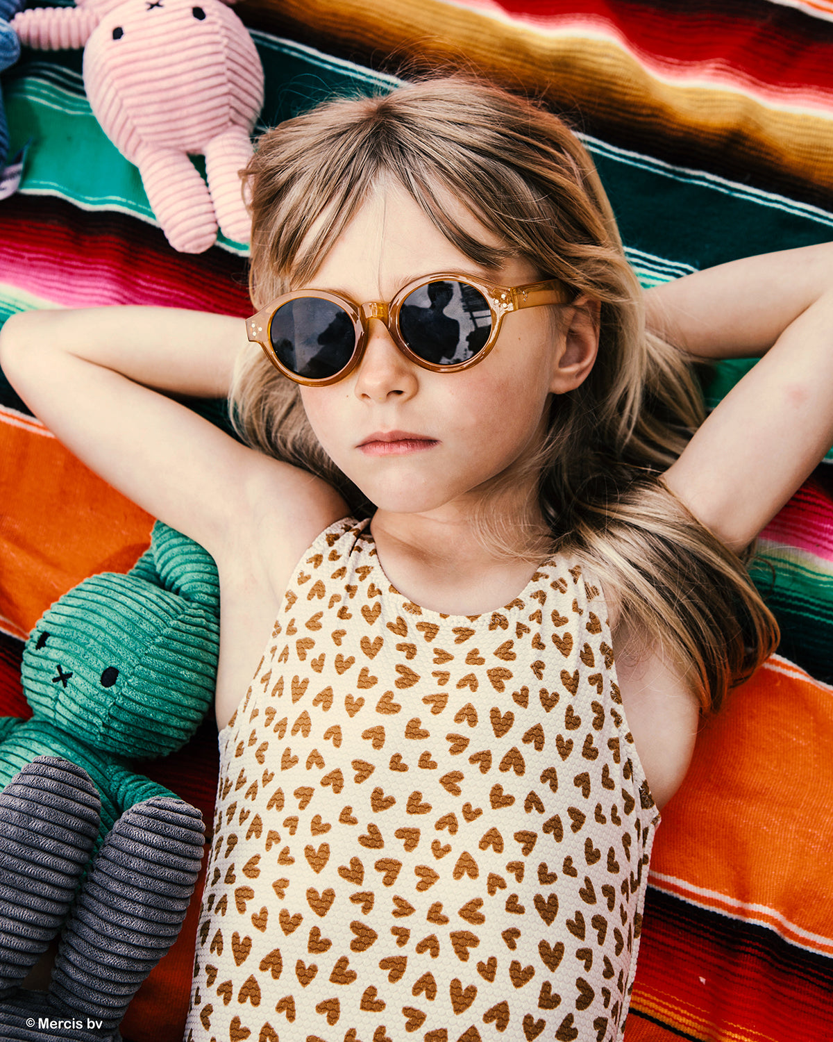 Child wearing sunglasses surrounded by colorful plush toys on a striped blanket