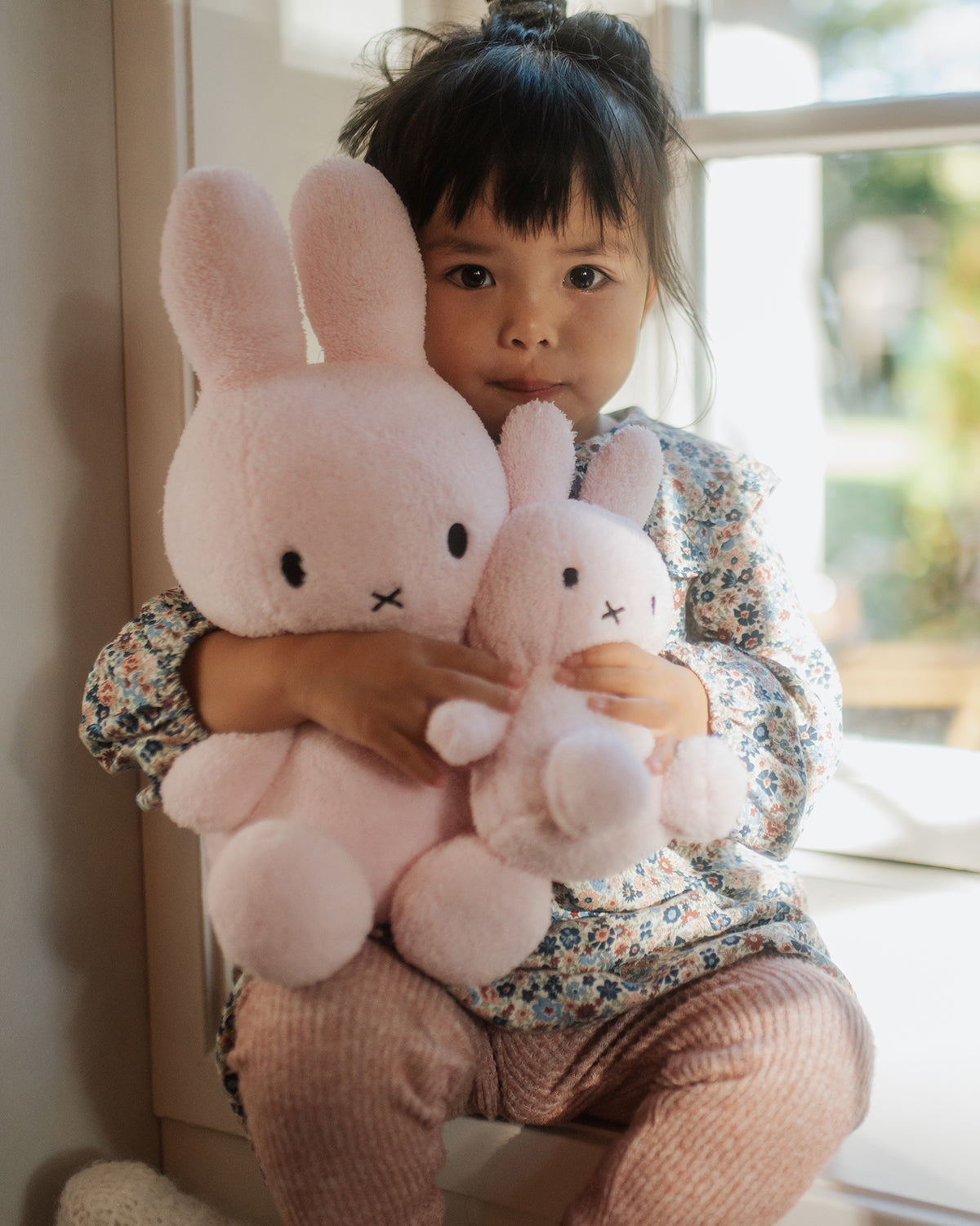Child holding two pink plush toys in a room with a window in the background