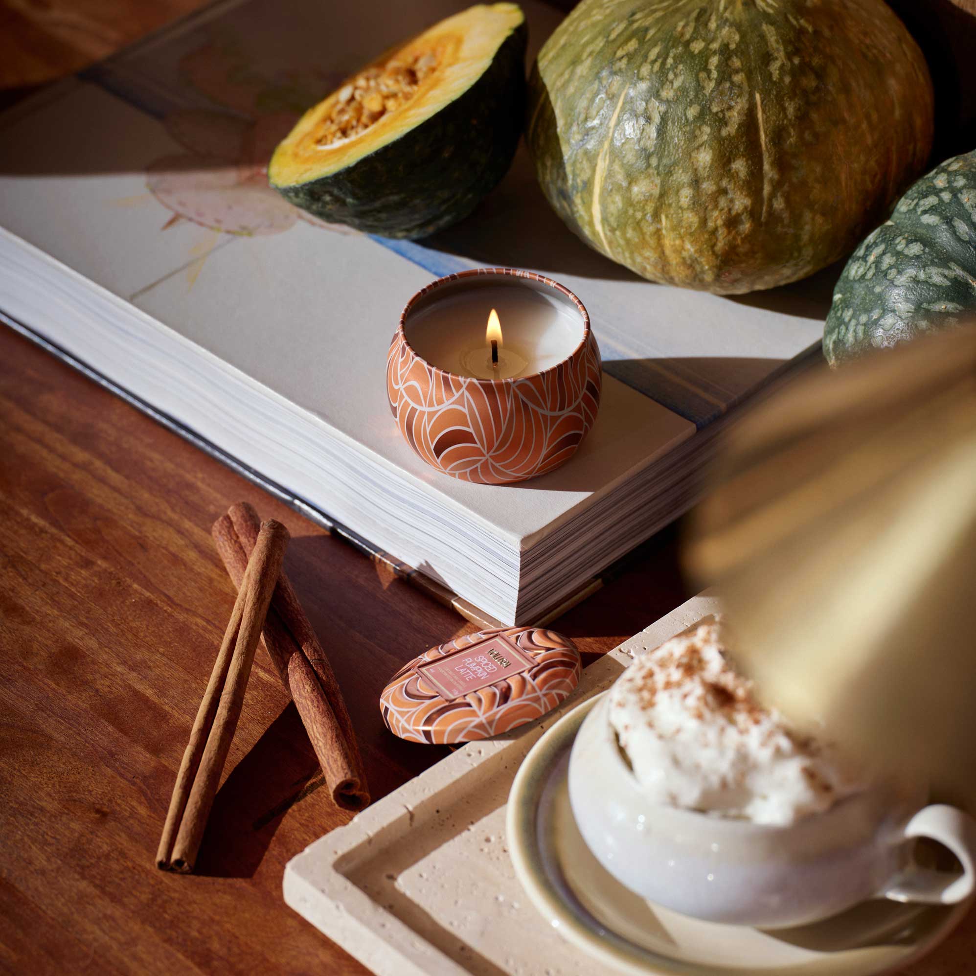 Candle burning on a book with pumpkins, cinnamon sticks, and a cup of coffee in the background.