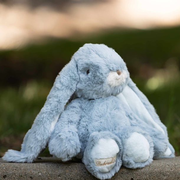 A light blue plush bunny stuffed animal sitting outdoors on a stone surface with a blurred green and brown background.