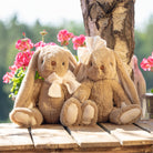Two plush bunnies with bows sitting on a wooden surface with flowers and a tree in the background.