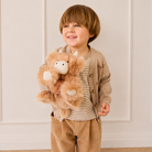 Soft brown plush Highland cow toy with white hooves and horns, held by a child in a striped shirt and beige cardigan, lifestyle front view.