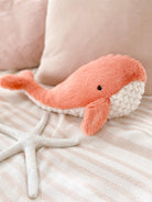 Stuffed, pink whale toy with white underside, resting on a striped blanket with a white starfish.
