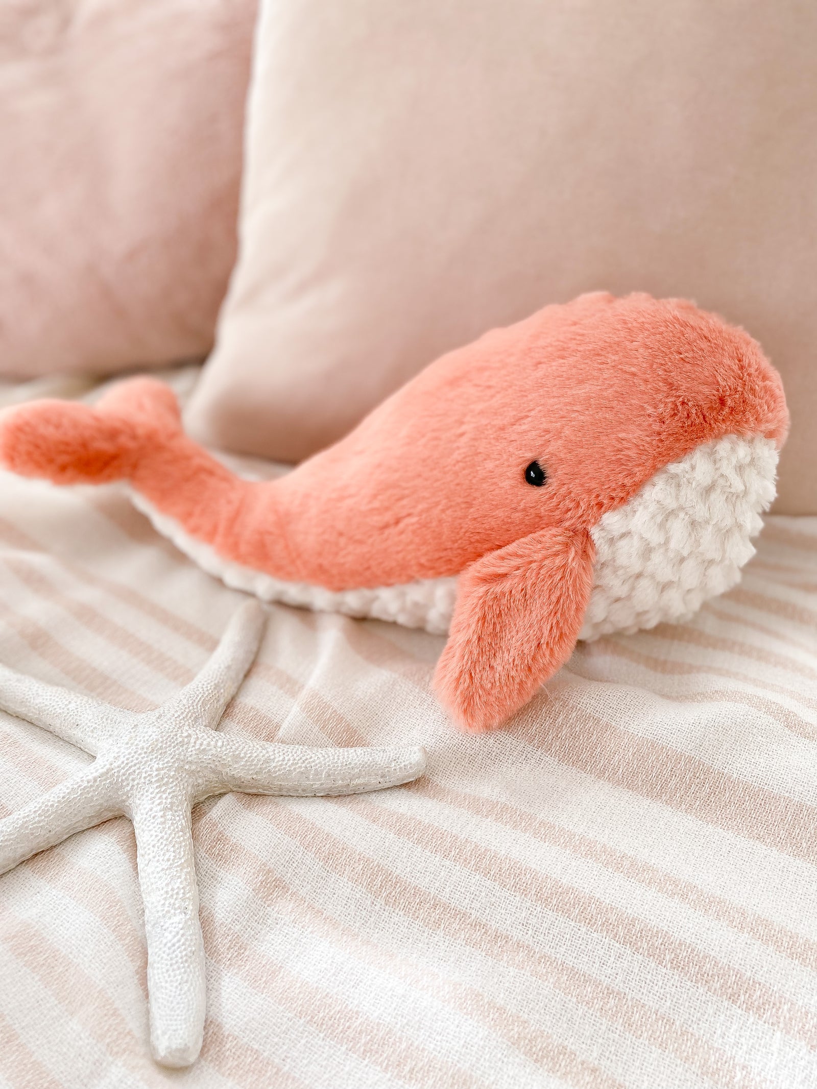 Stuffed, pink whale toy with white underside, resting on a striped blanket with a white starfish.
