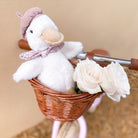 A white duck plush toy with a pink beret and floral scarf sits in a wicker bicycle basket with two white roses.