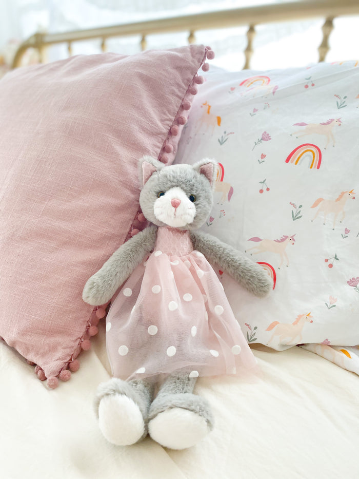 Stuffed gray cat toy wearing a pink dress with white polka dots, sitting on a bed with a pink pillow and unicorn-themed bedding.